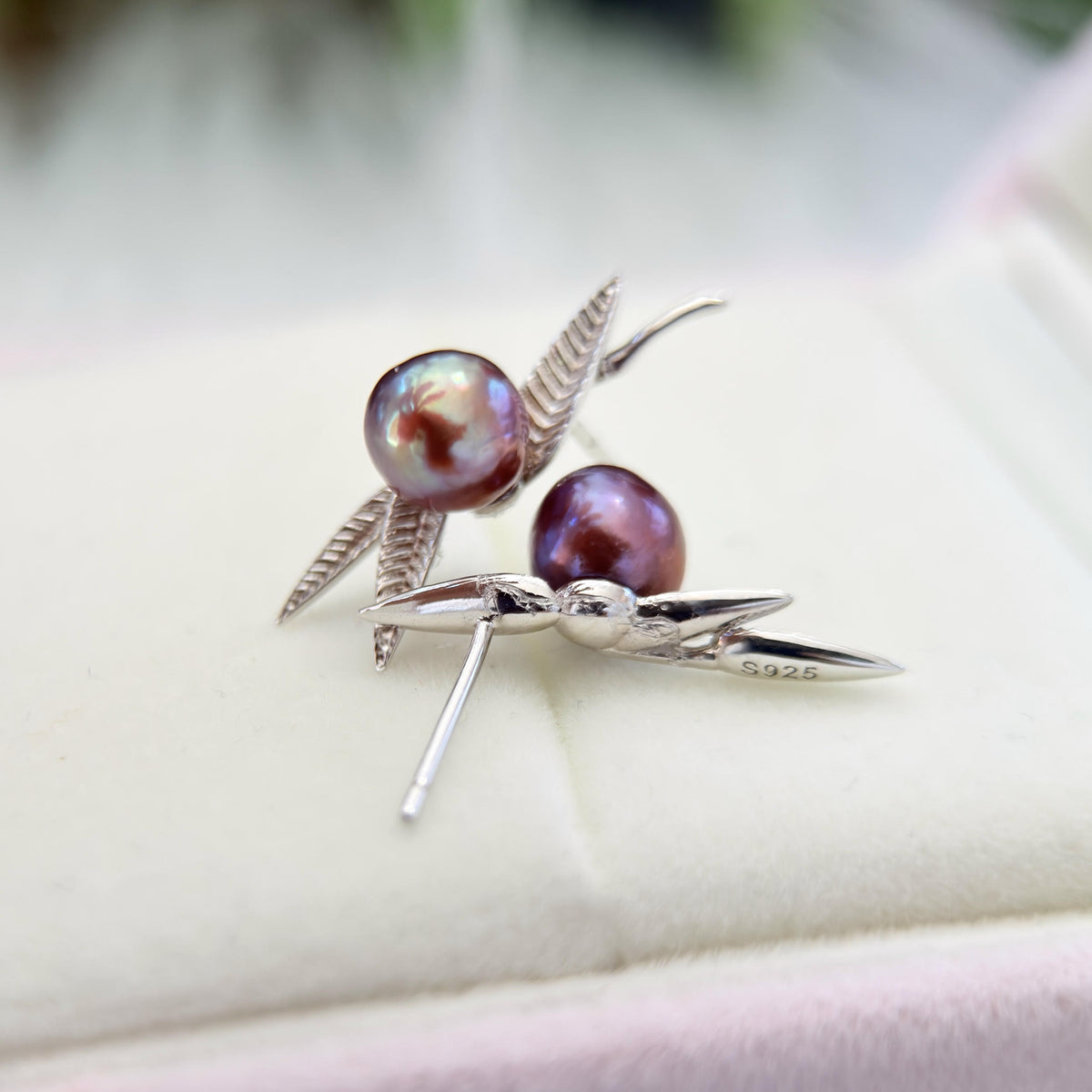 A pair of silver dragonfly stud earrings with iridescent purple pearls, displayed on a soft cream-colored jewelry box.