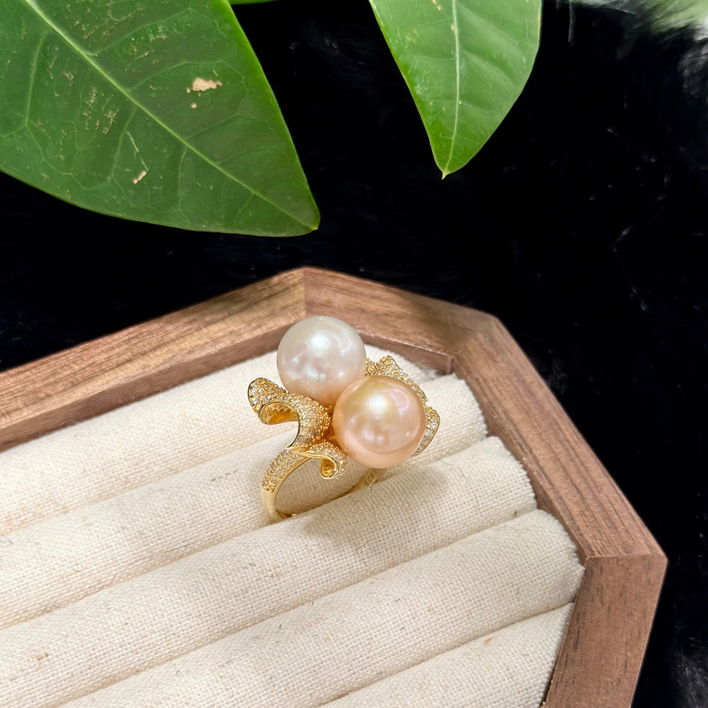 A gold ring featuring two large, round, lustrous pearls in soft pink and cream tones, displayed on a cream-colored jewelry tray inside a wooden box with green leaves in the background.