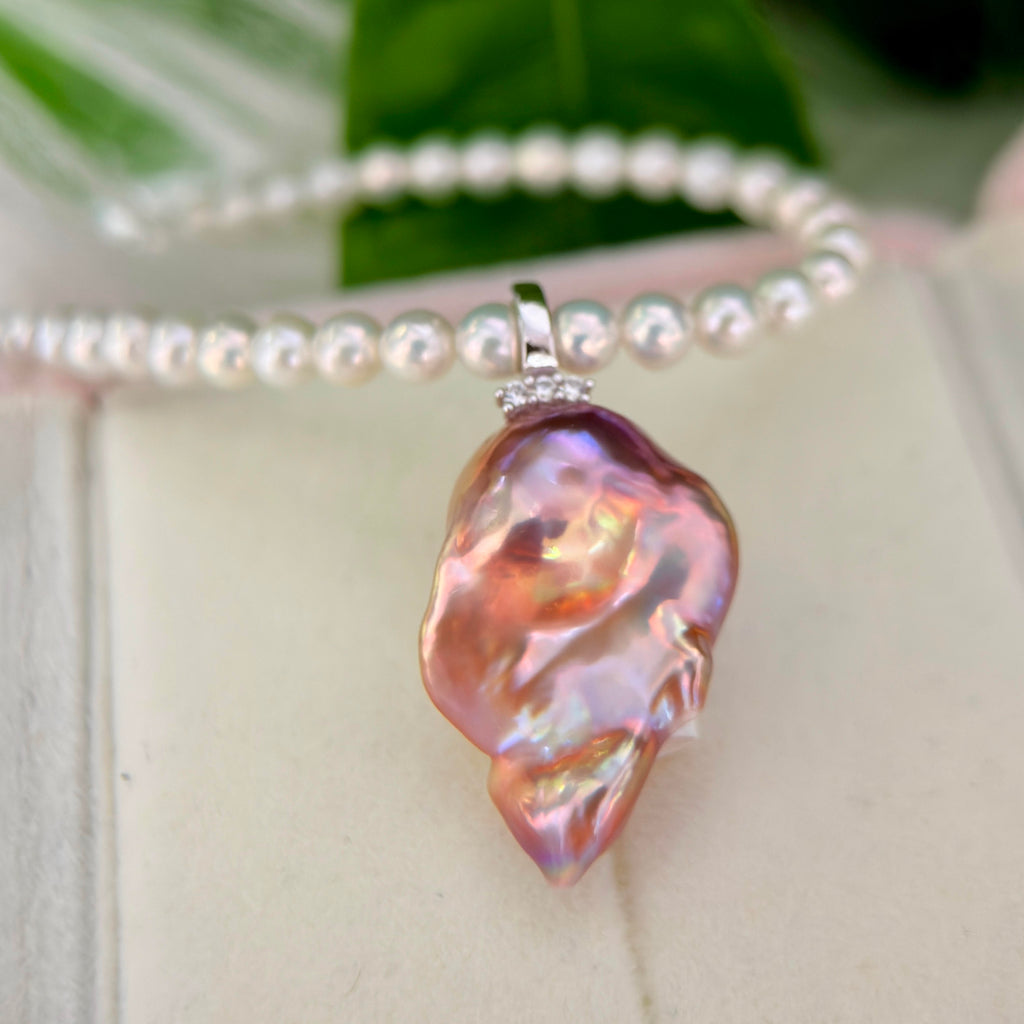 A close-up of a baroque pearl pendant necklace with a pink, iridescent finish and a silver bezel set with small clear pearls, displayed on a light cream surface with a pearl bracelet in the background.