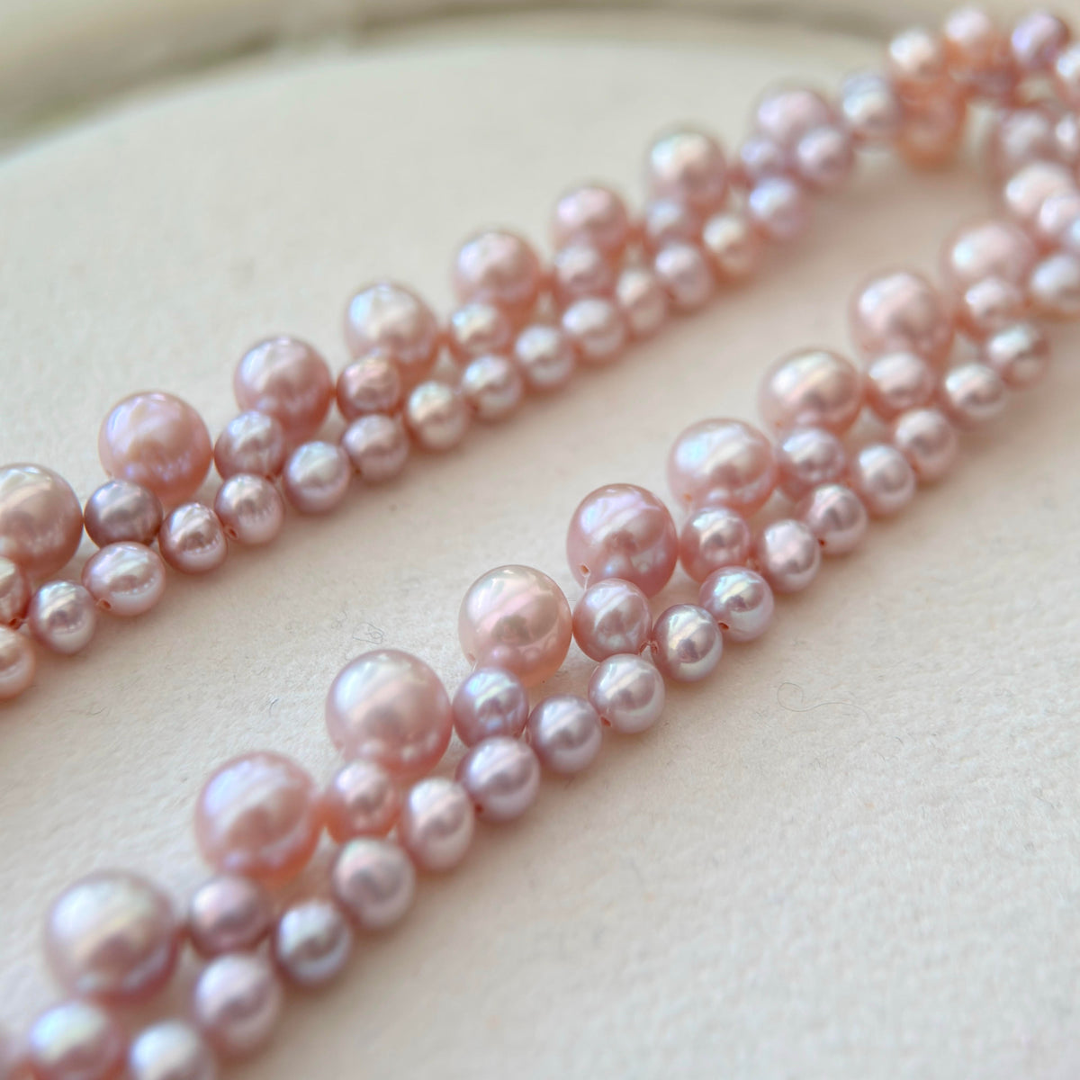 Two strands of freshwater pearl beads in a mix of sizes, featuring a soft pink and peach iridescent luster, arranged diagonally on a light beige textured surface.