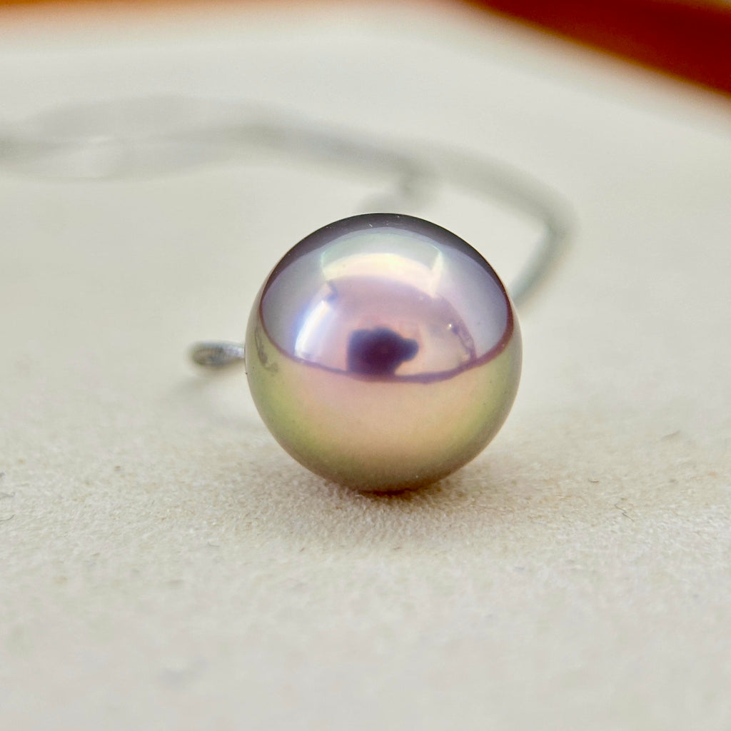 A single round freshwater pearl pendant with iridescent lavender and green tones, mounted on a silver post, shown close-up on a light beige textured surface.