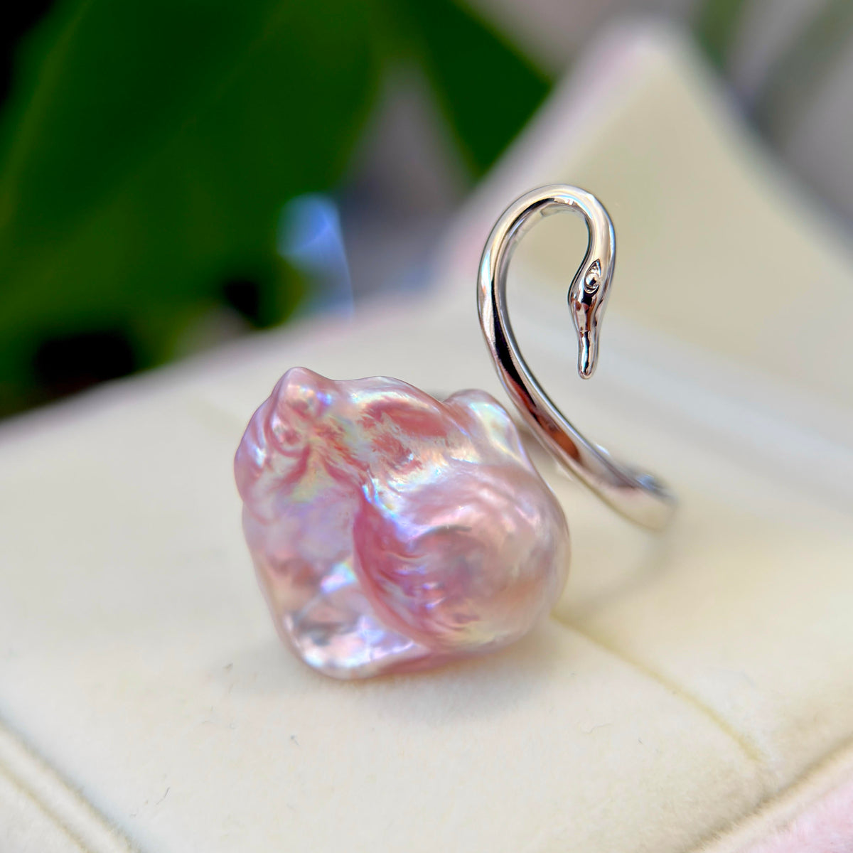 A silver ring shaped like a swan with a large, lustrous pink baroque pearl, displayed on a cream-colored ring box with a blurred green background.