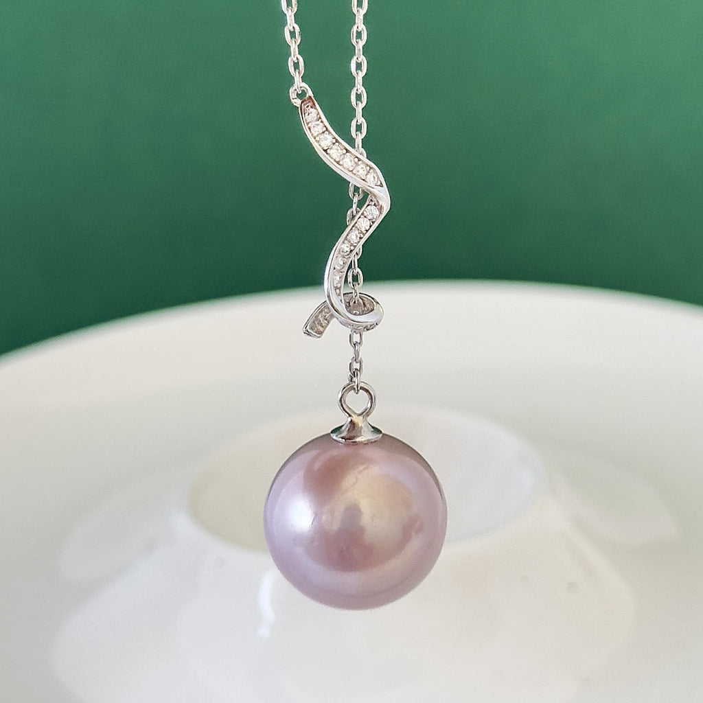 A silver pendant necklace featuring a wavy, diamond-accented chain and a single large, lustrous pink pearl drop, shown against a dark green background and white dish.