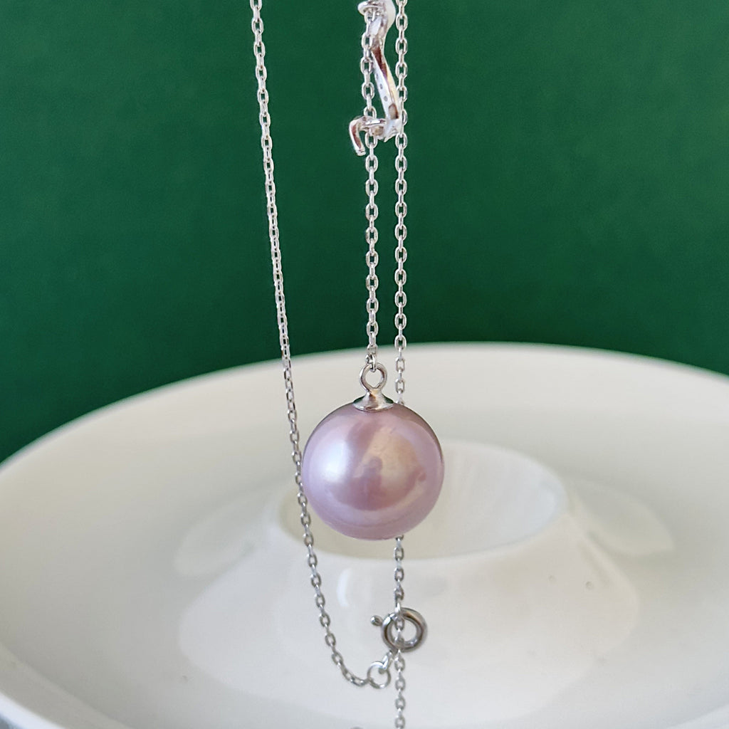 A silver chain necklace featuring a large, lustrous pinkish-purple freshwater pearl pendant, displayed on a white ceramic dish against a dark green background.