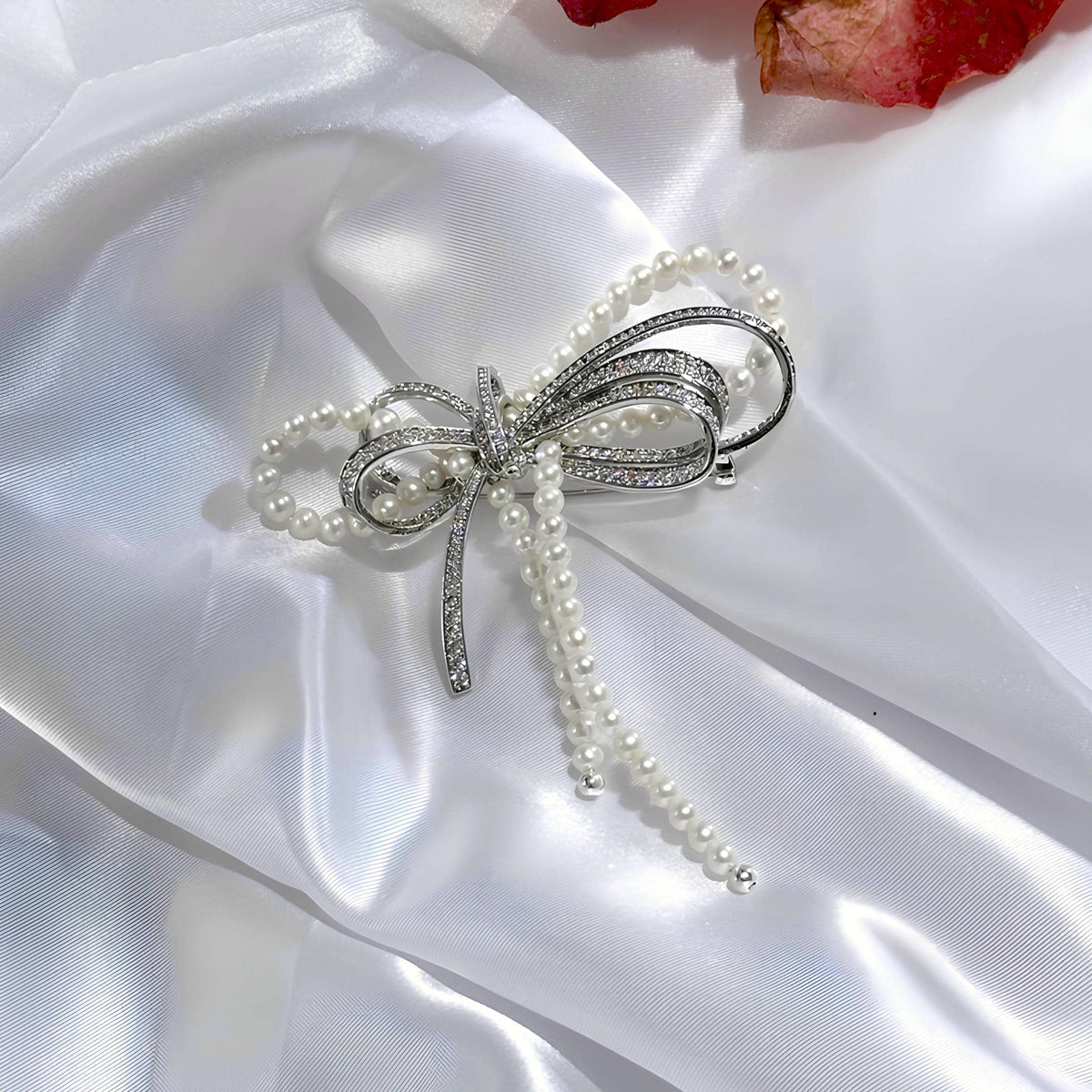 A silver bow-shaped brooch embellished with clear rhinestones and adorned with dangling strands of white faux pearls, displayed on a draped white satin fabric background.