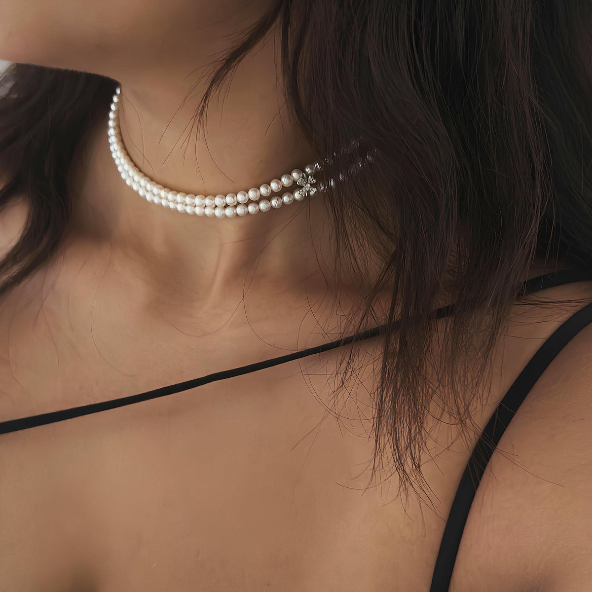 A close-up of a model wearing a double-strand pearl choker necklace with small clear rhinestone accents at the front, paired with a black spaghetti-strap top.
